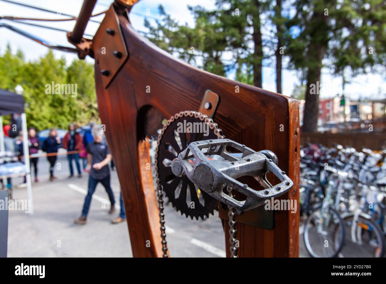 A close up view on the details of a wood bike stood on end. Metal chain and pedal are seen in foreground, parked bicycles and blurry people in backgro Stock Photo