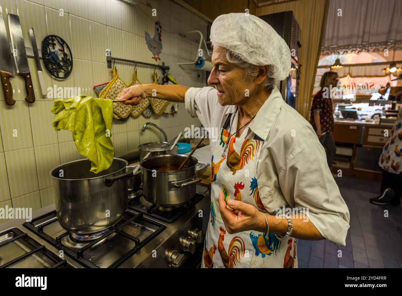 In the kitchen of the Le Sfogline pasta workshop, an employee stirs two pots on the stove with a green cloth; through the doorway in the background, the shop's sign is visible. Le Sfogline, at Via Belvedere 7/b, is run by the sisters Monica and Daniela, who took over the business from their mother, Renata Zappoli, and make tortellini, tagliatelle and gramigna, among other fresh pasta. Via Belvedere, Bologna, Emilia-Romagna, Italy. Stock Photo
