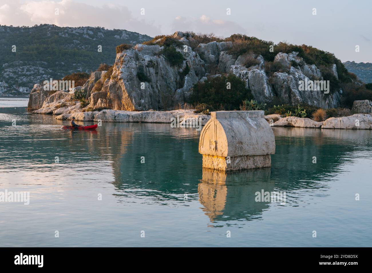 Stone sarcophagus from ancient Simena Kaleköy city Turkey, sits partially submerged in clear waters of Mediterranean surrounded by rugged hills listed Stock Photo