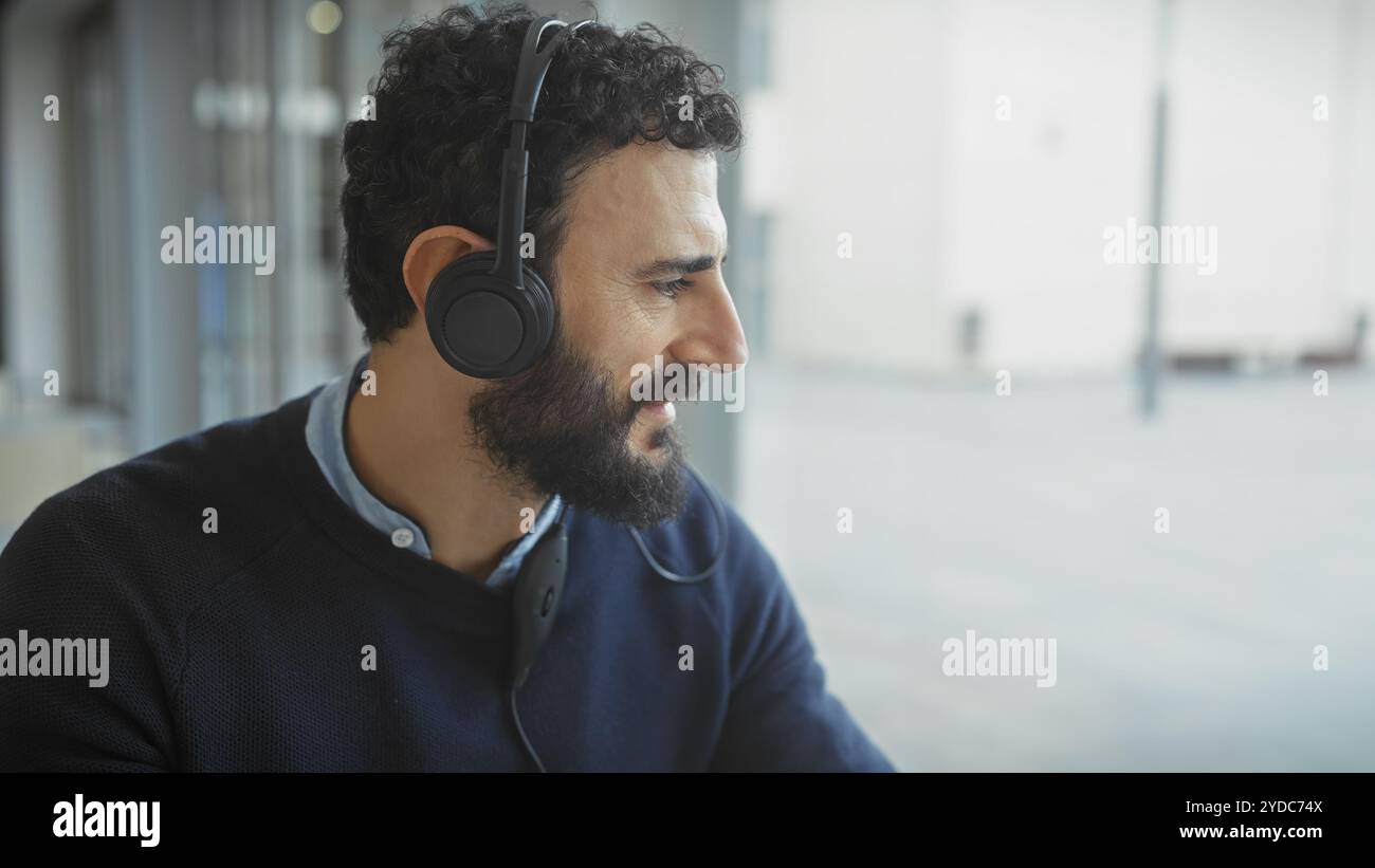 A bearded man wearing headphones in a modern office setting, looking away thoughtfully. Stock Photo