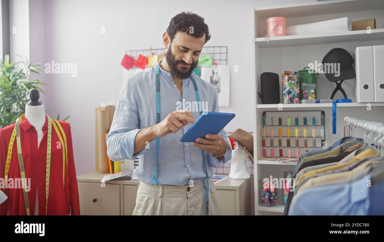 A bearded tailor in a workshop using a tablet amongst mannequins, fabric, and colorful threads. Stock Photo