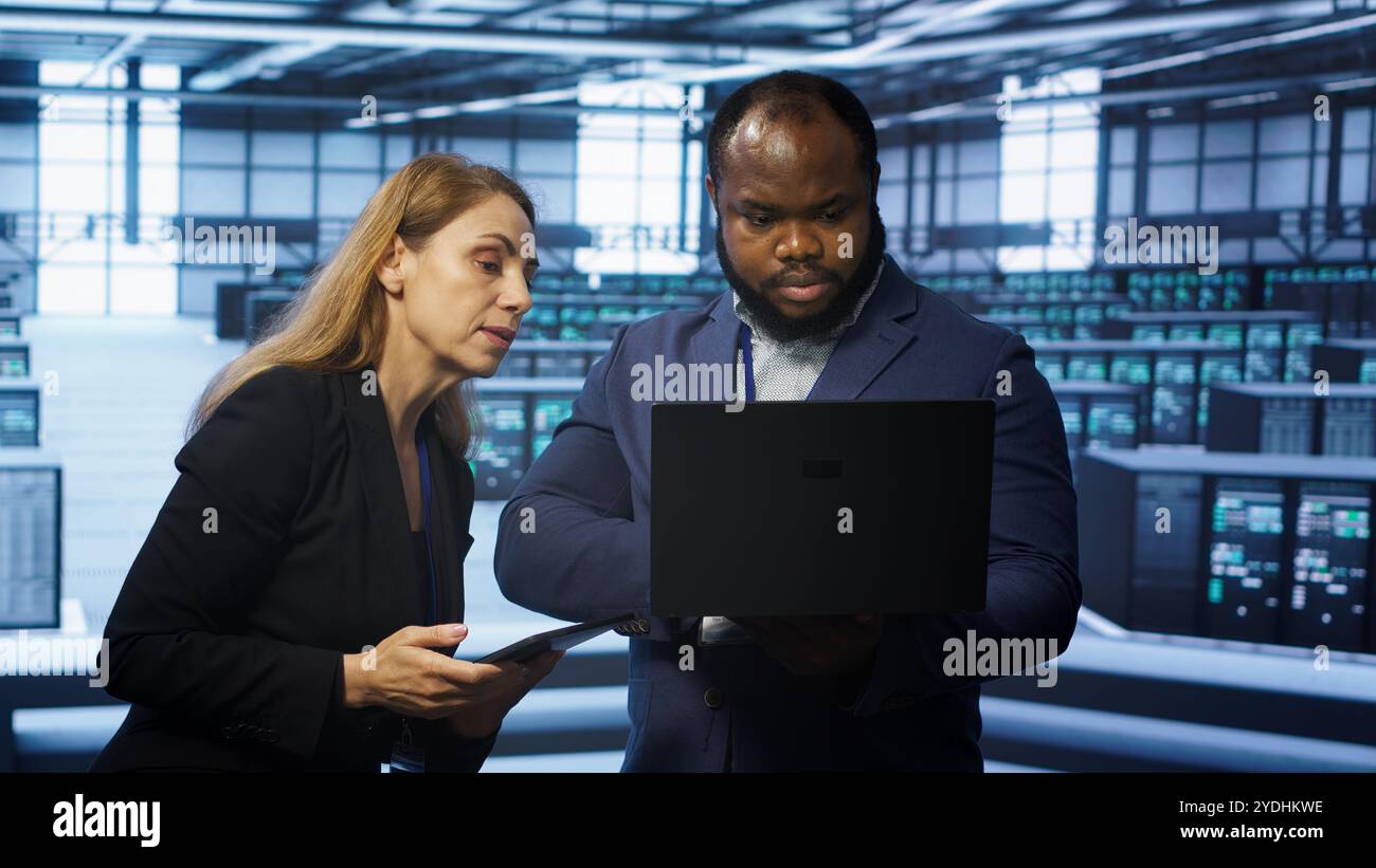 System administrators in data center overseeing server operations using notebook and tablet. Colleagues brainstorming in server hub, ensuring optimal performance and data integrity, camera A Stock Photo