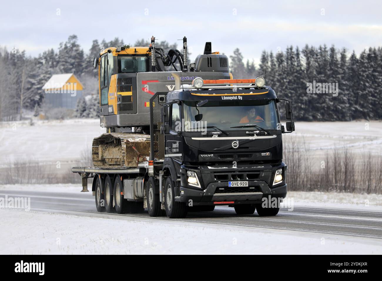 Black Volvo FMX truck Lonnqvist Oy hauls Volvo excavator on flatbed trailer along highway 52 on a day of winter. Salo, Finland. December 27, 2021 Stock Photo