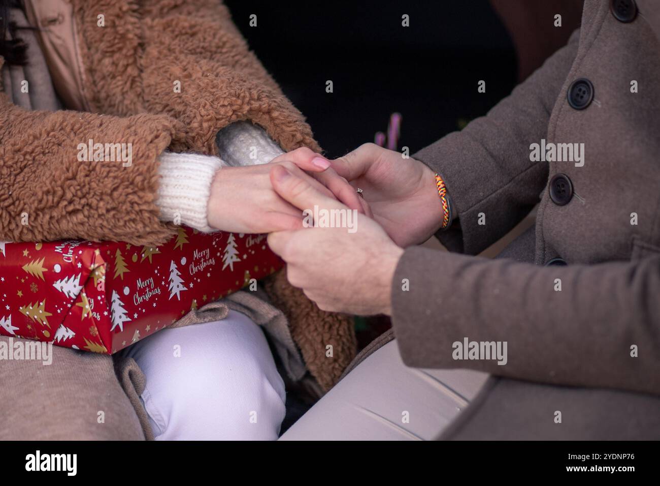 A couple is enjoying a warm and intimate moment together, sitting close while holding hands near a beautifully wrapped holiday gift, creating a memora Stock Photo