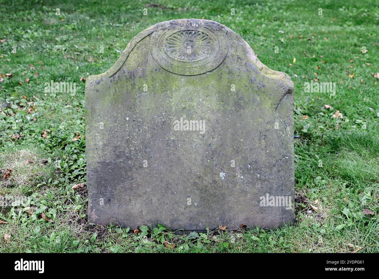 A closeup of a very old tombstone. The stone is blank except for a design at the top. The stone is from the 18th or 19th century. Grass surrounds it. Stock Photo