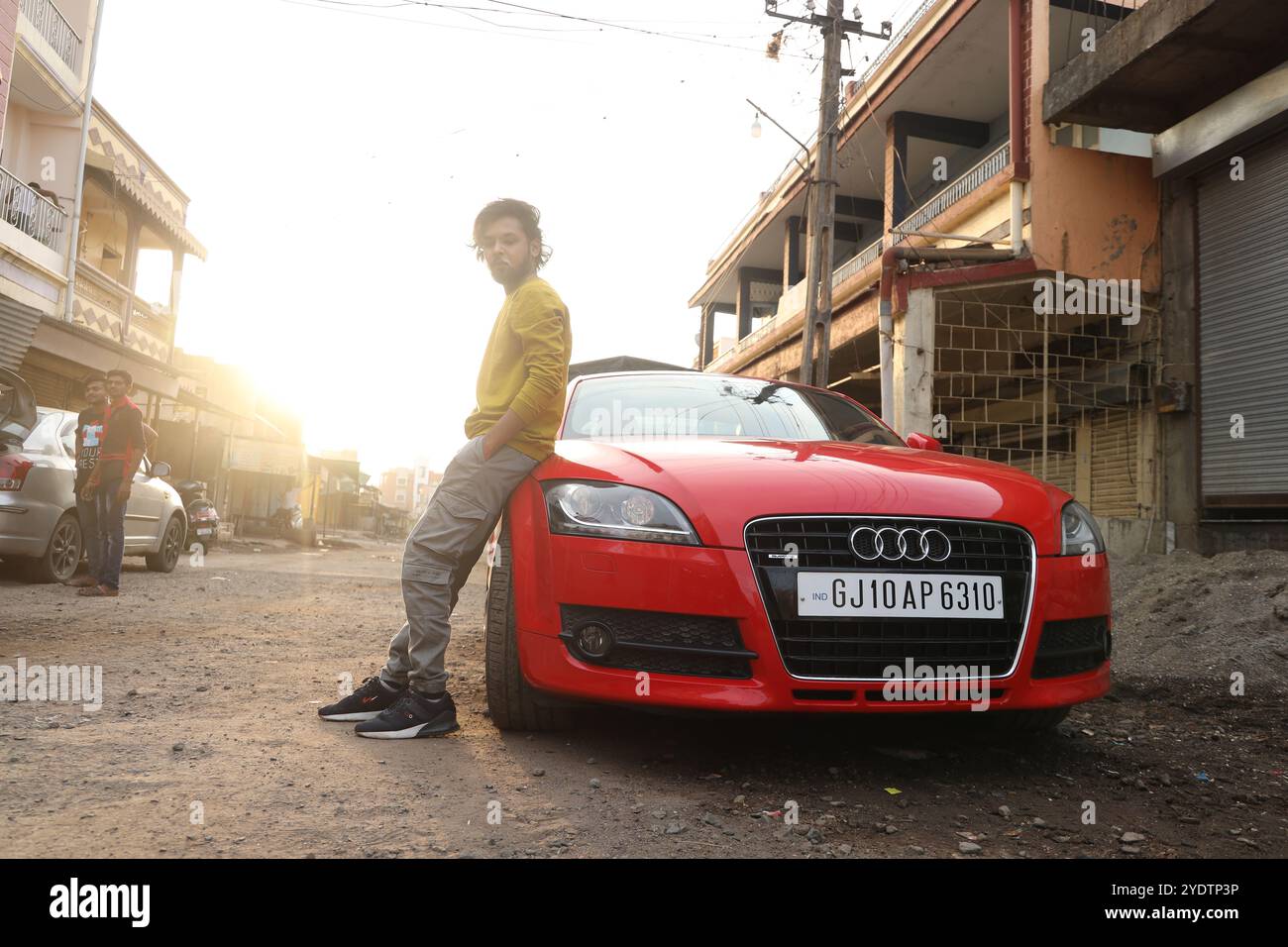 A confident man in a mustard yellow shirt sits casually inside a sleek red Audi TT with black rims, parked against a rugged industrial backdrop. The open car door and his intense gaze add to the bold, urban vibe of the scene, blending luxury with an edgy, street-style aesthetic. Stock Photo
