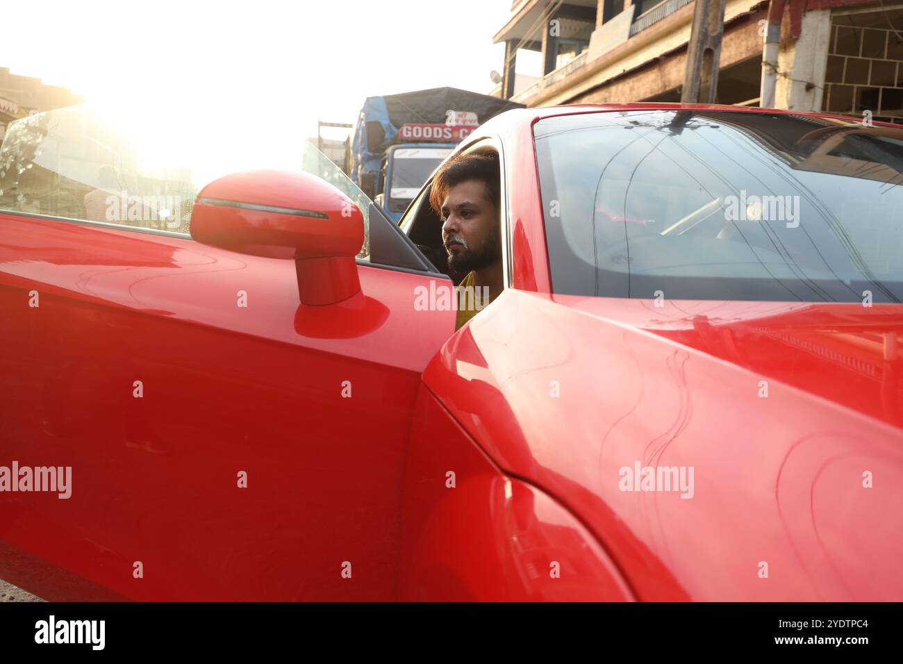 A confident man in a mustard yellow shirt sits casually inside a sleek red Audi TT with black rims, parked against a rugged industrial backdrop. The open car door and his intense gaze add to the bold, urban vibe of the scene, blending luxury with an edgy, street-style aesthetic. Stock Photo