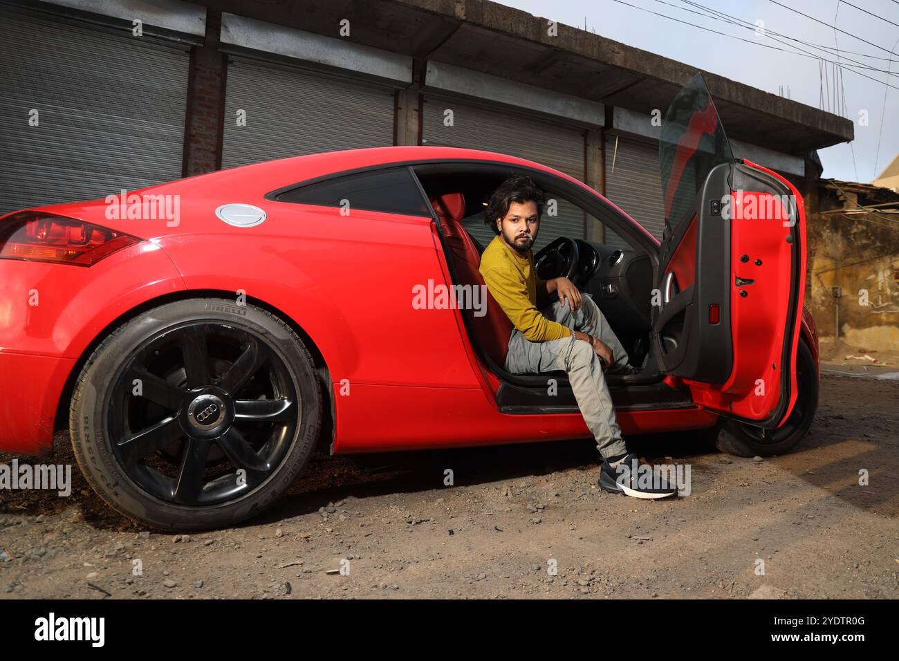 A confident man in a mustard yellow shirt sits casually inside a sleek red Audi TT with black rims, parked against a rugged industrial backdrop. The open car door and his intense gaze add to the bold, urban vibe of the scene, blending luxury with an edgy, street-style aesthetic. Stock Photo