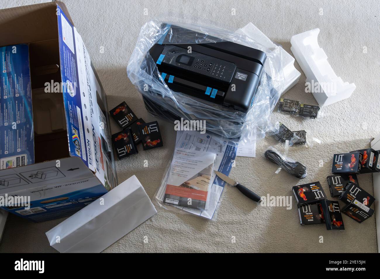 A new Epson home printer being unwrapped on a carpet floor in a UK house with waste packaging, printer paper, ink cartridges and plastic wrapping Stock Photo