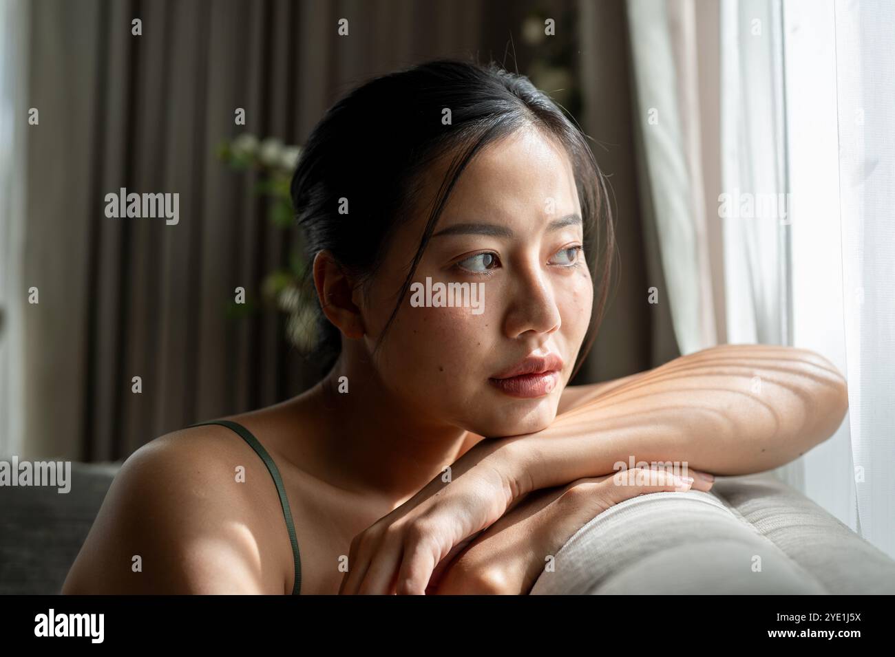 A beautiful, thoughtful Asian woman is sitting on a sofa at home ...