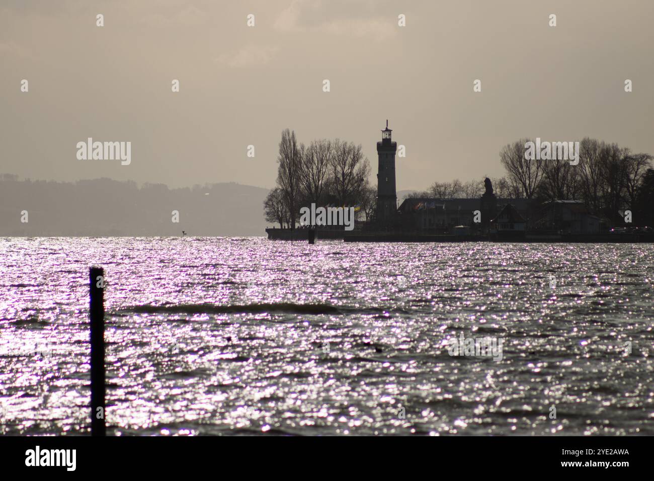 Silhouette of the Lindau lighthouse by Lake Constance, with shimmering waters and a moody, cloudy sky in the background. Stock Photo