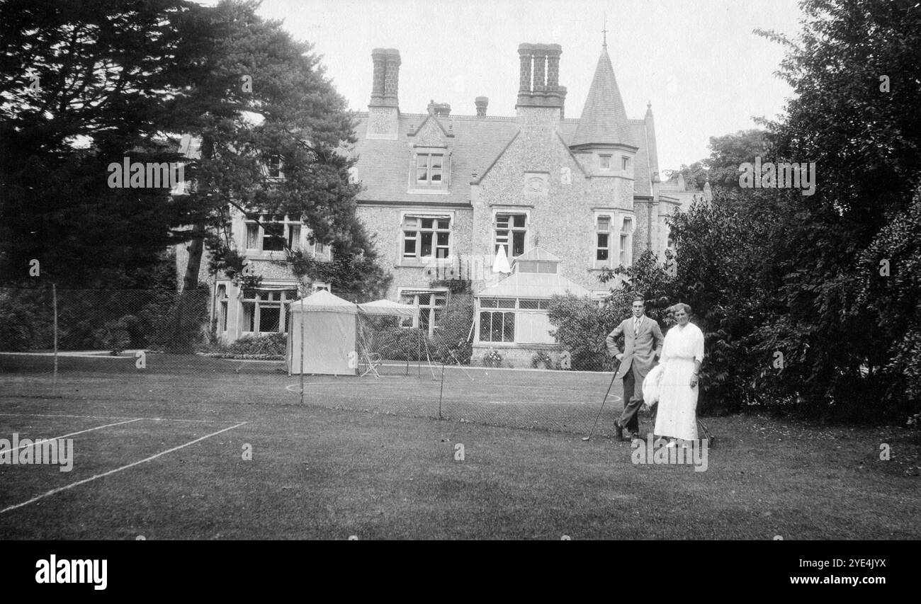 West Sussex, England. c.1913 – A young man and a woman, believed to be relatives of the Henty family, are posing for a photograph in the tennis court within the grounds of Ferring Grange, in Ferring, a coastal village in West Sussex. The man is holding a golf club. This estate was the home of Edwin Henty, J.P, D.L., F.S.A. (1844 – 1916), who had served as High Sheriff of Sussex. In 1924, the house was converted into a fashionable hotel, visited by many celebrities including Edward, the Prince of Wales. The house was destroyed by fire in October 1946. Stock Photo