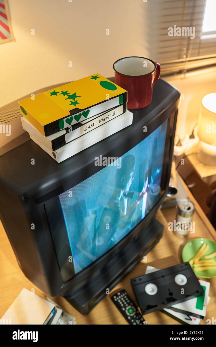 Stacked VHS tapes with old CRT television and assortment of vintage media equipment surrounding it. Includes coffee cup and blinds in the dimly lit scene Stock Photo