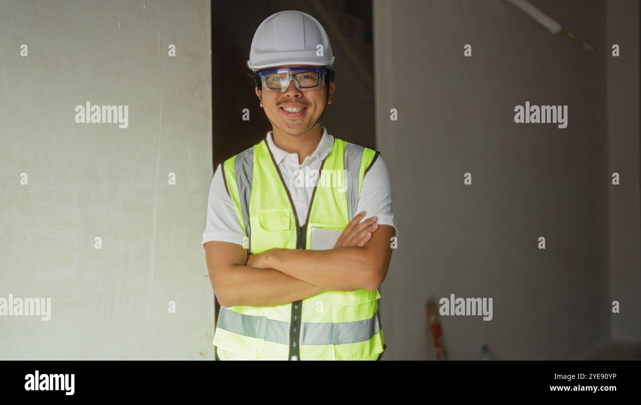 Young man wearing safety gear standing indoors with arms crossed at a construction site Stock Photo