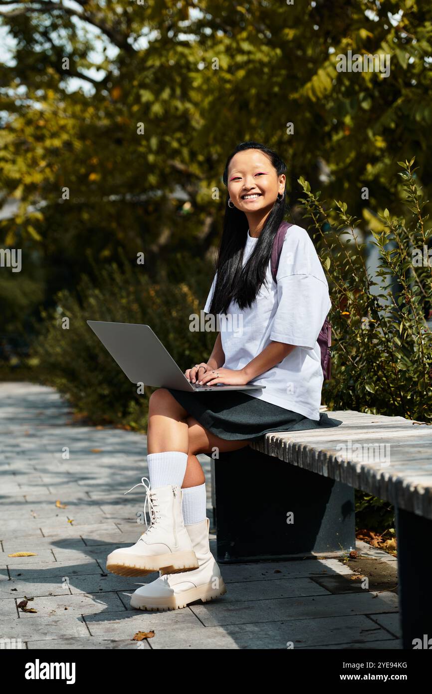 A cheerful young Asian girl sits on a bench, focused on her laptop ...