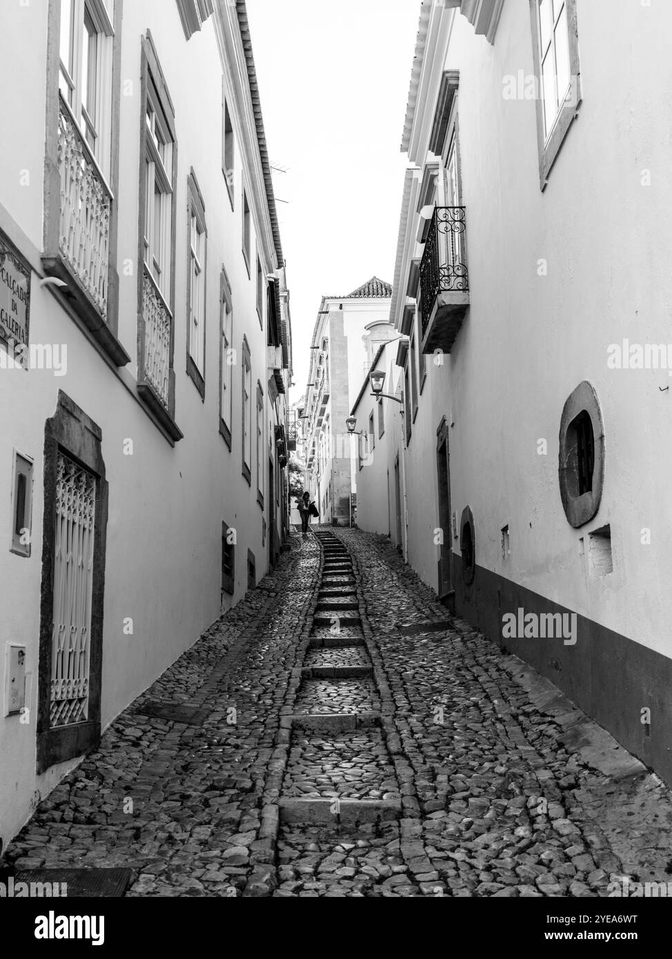 View through cobblestone street of person walking along side the whitewashed buildings in the Moorish town of Tavira, a small city on the Algarve C... Stock Photo