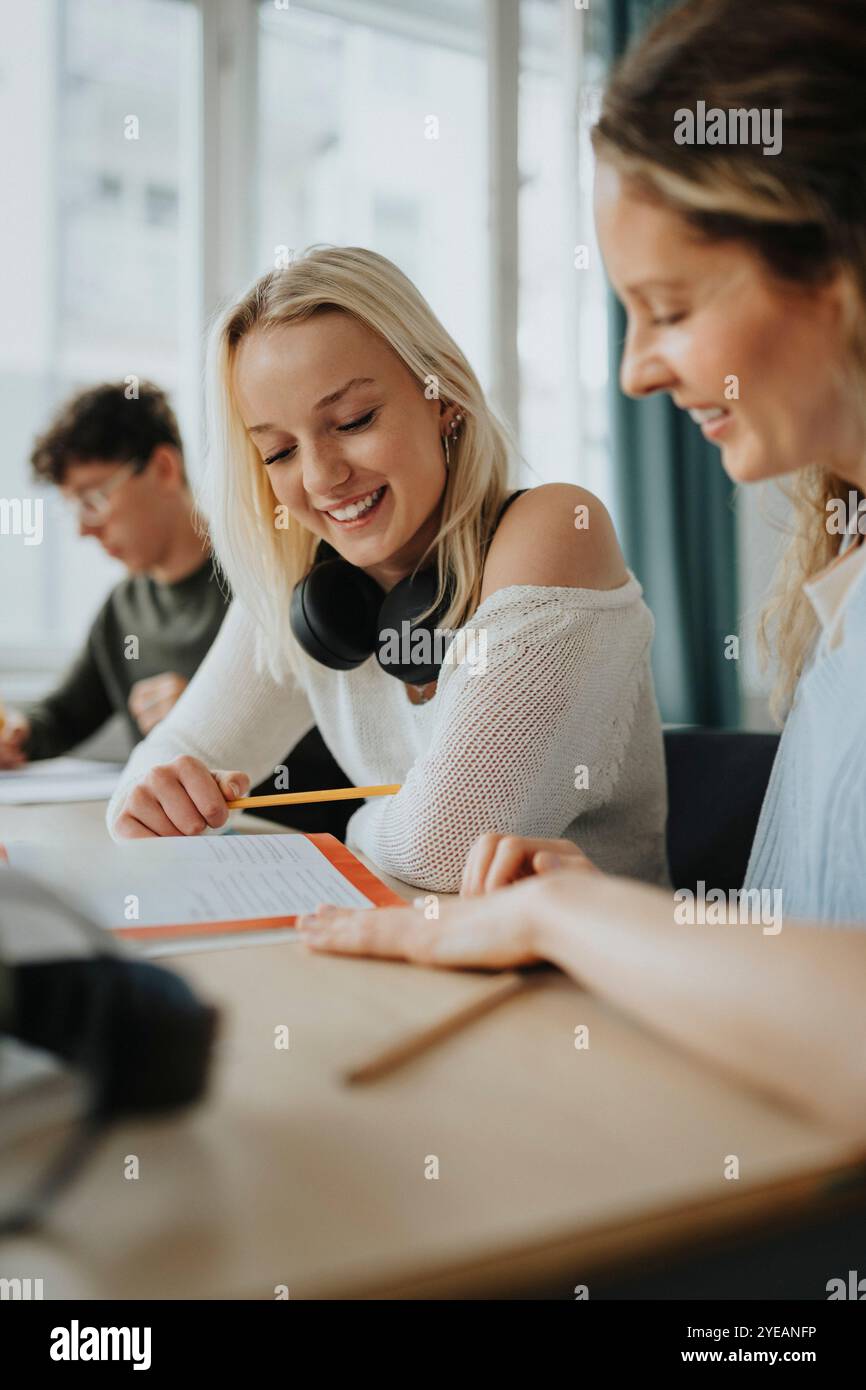 Happy blond girl reading book by female teacher sitting at desk Stock ...