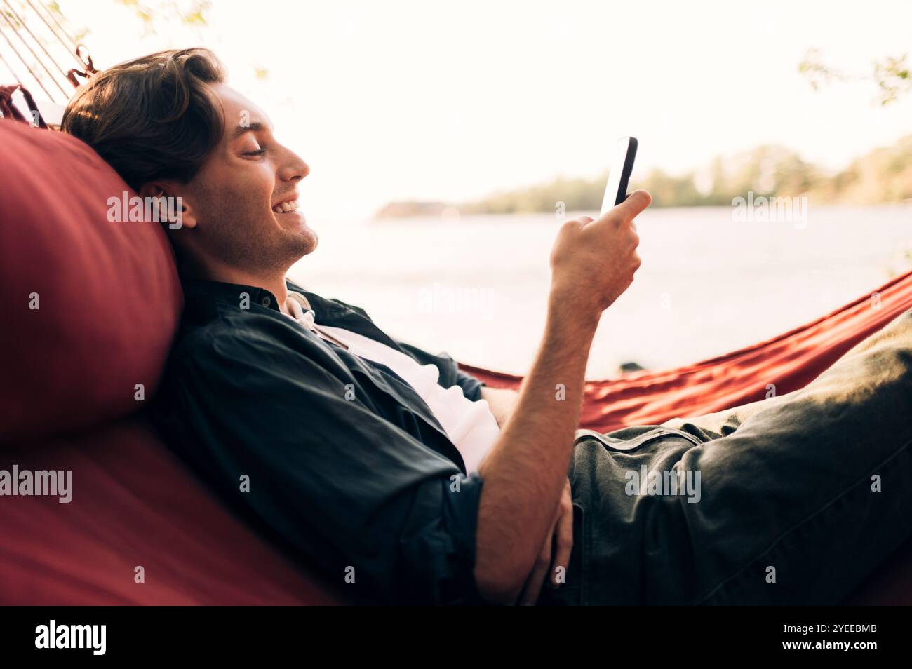Side view of smiling man using smart phone while lying down in hammock Stock Photo