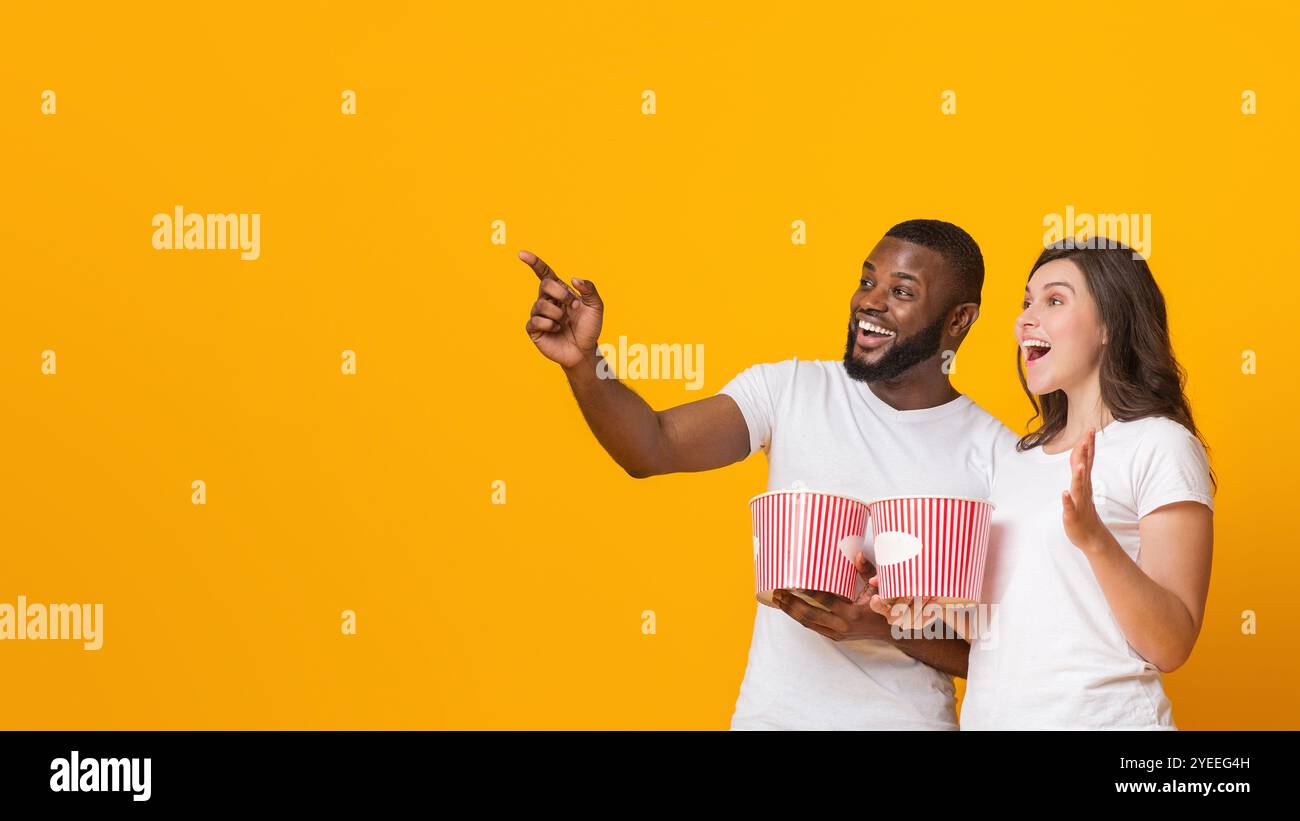 Excited interracial couple holding popcorn backets and pointing at copy space Stock Photo