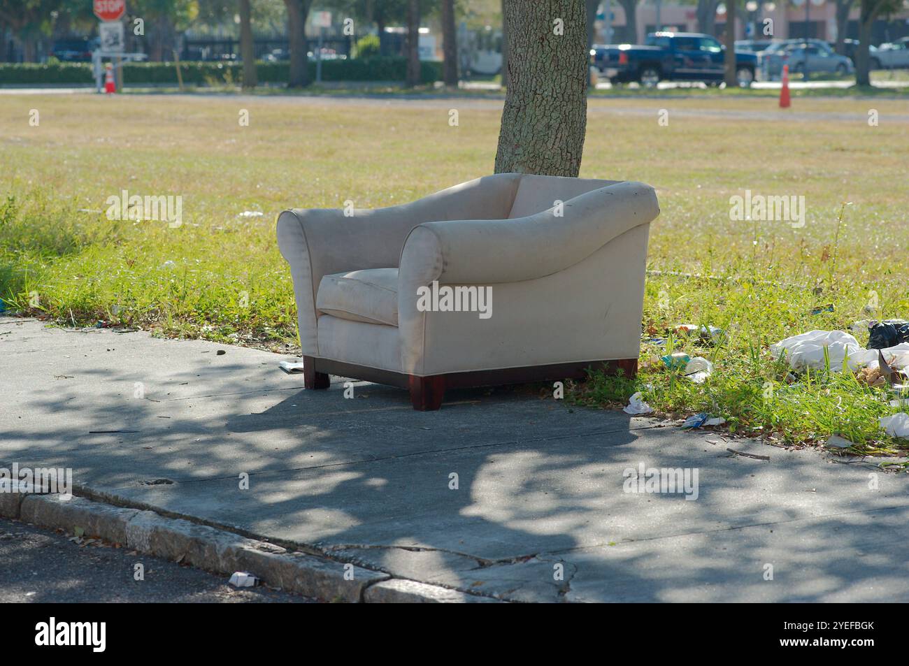 Isolated light tan arm chair discarded on sidewalk in sunshine and shade, Tree trunk and green grass in background and street intersection. Trash besi Stock Photo