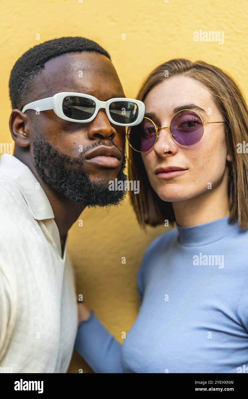 Vertical close-up photo of a stylish multiracial couple wearing sunglasses looking at camera together against yellow background Stock Photo
