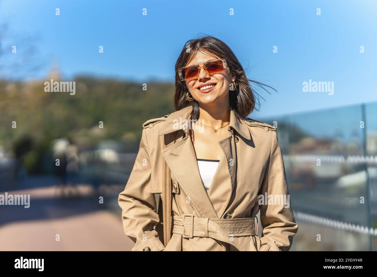 A woman wearing a tan coat and sunglasses is smiling and walking down a street. Concept of happiness and leisure, as the woman is enjoying her time ou Stock Photo