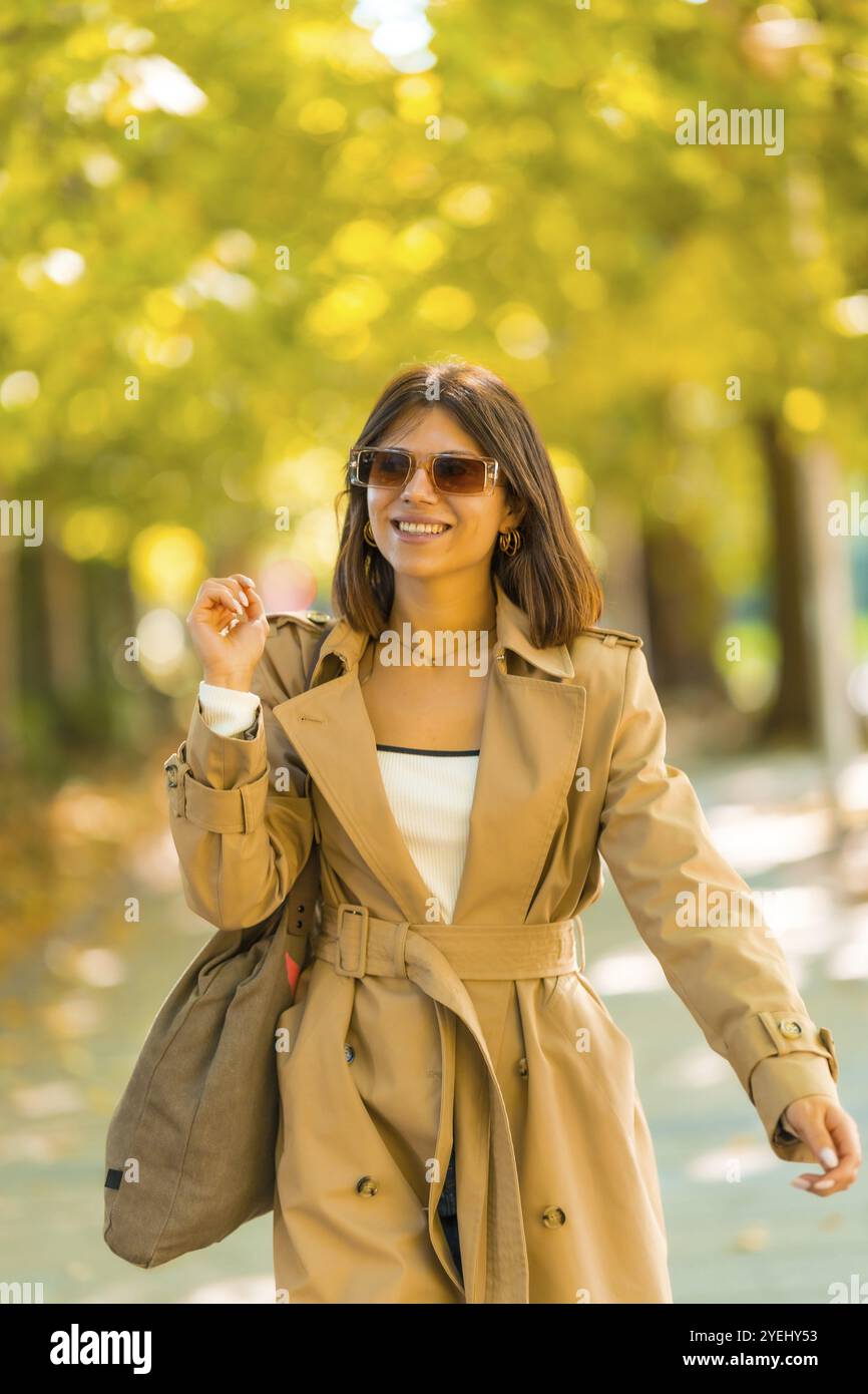 A woman wearing a tan trench coat and sunglasses walks down a sidewalk. She is smiling and holding a handbag. Autumn or winter concept in the city Stock Photo
