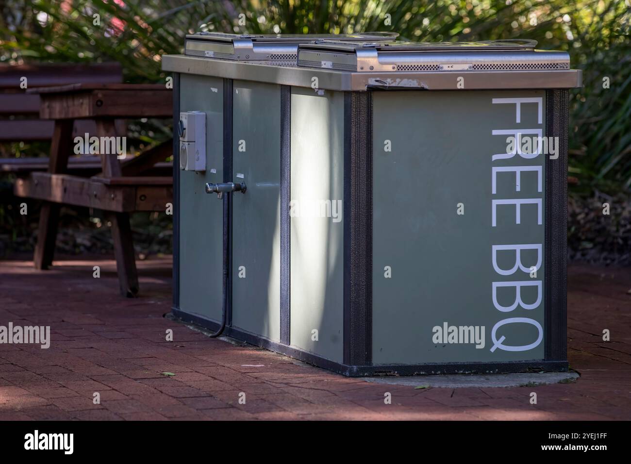 This photo captures a public 'Free BBQ' unit in a park setting, with a wooden bench and green vegetation in the background. Stock Photo