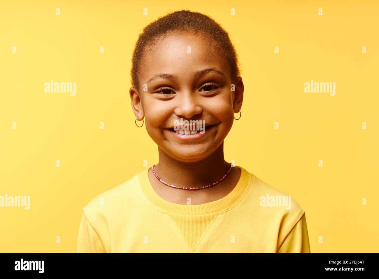 Portrait of cute cheerful girl of African American ethnicity wearing colorful beaded choker smiling at camera while standing against yellow background in studio, copy space Stock Photo