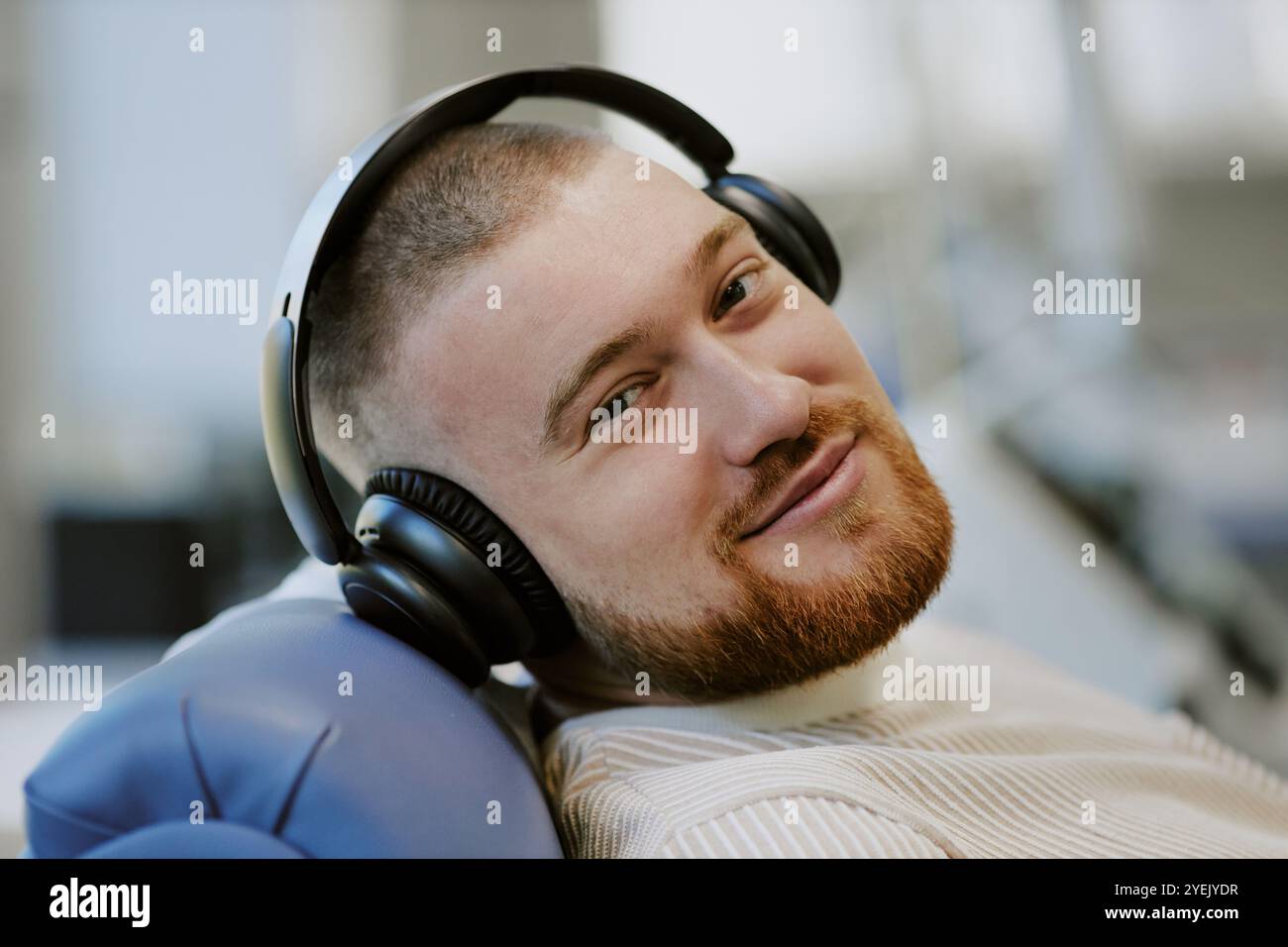 Man with headphones sitting in a relaxed position while enjoying music and smiling. He is looking straight at the camera with a friendly expression, seated indoors Stock Photo