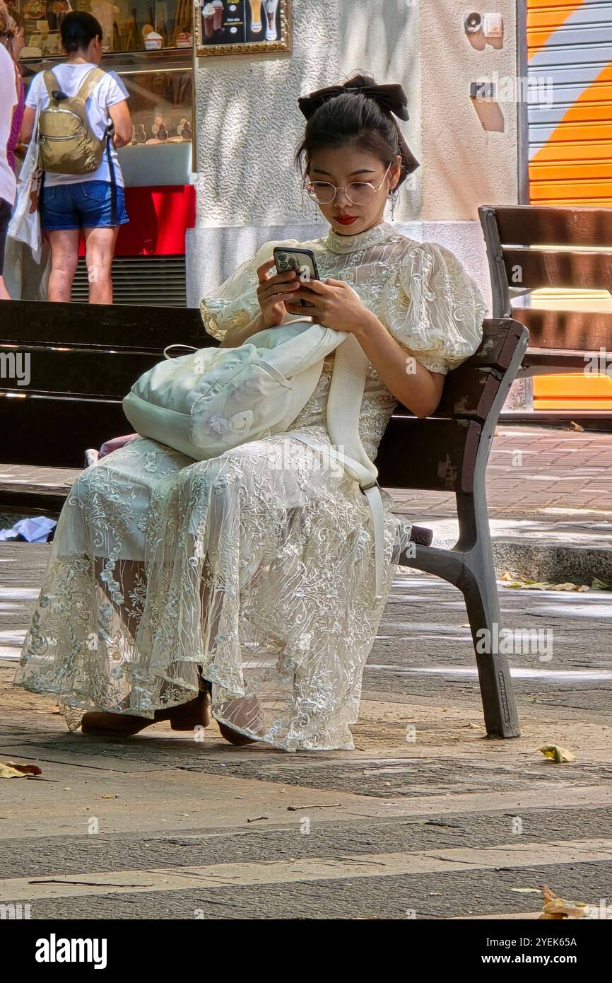 An Asian woman wearing a white dress sitting on a bench at Avinguda de Gaudí in Barcelona, Spain. - Smartphone Captured Stock Image
