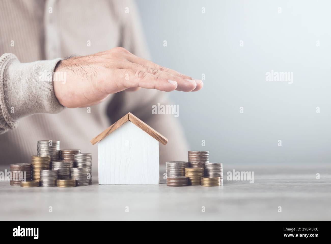 A businessman making a protective hand gesture over a stack of coins and a house model. Image represents the concept of asset protection, emphasizing Stock Photo