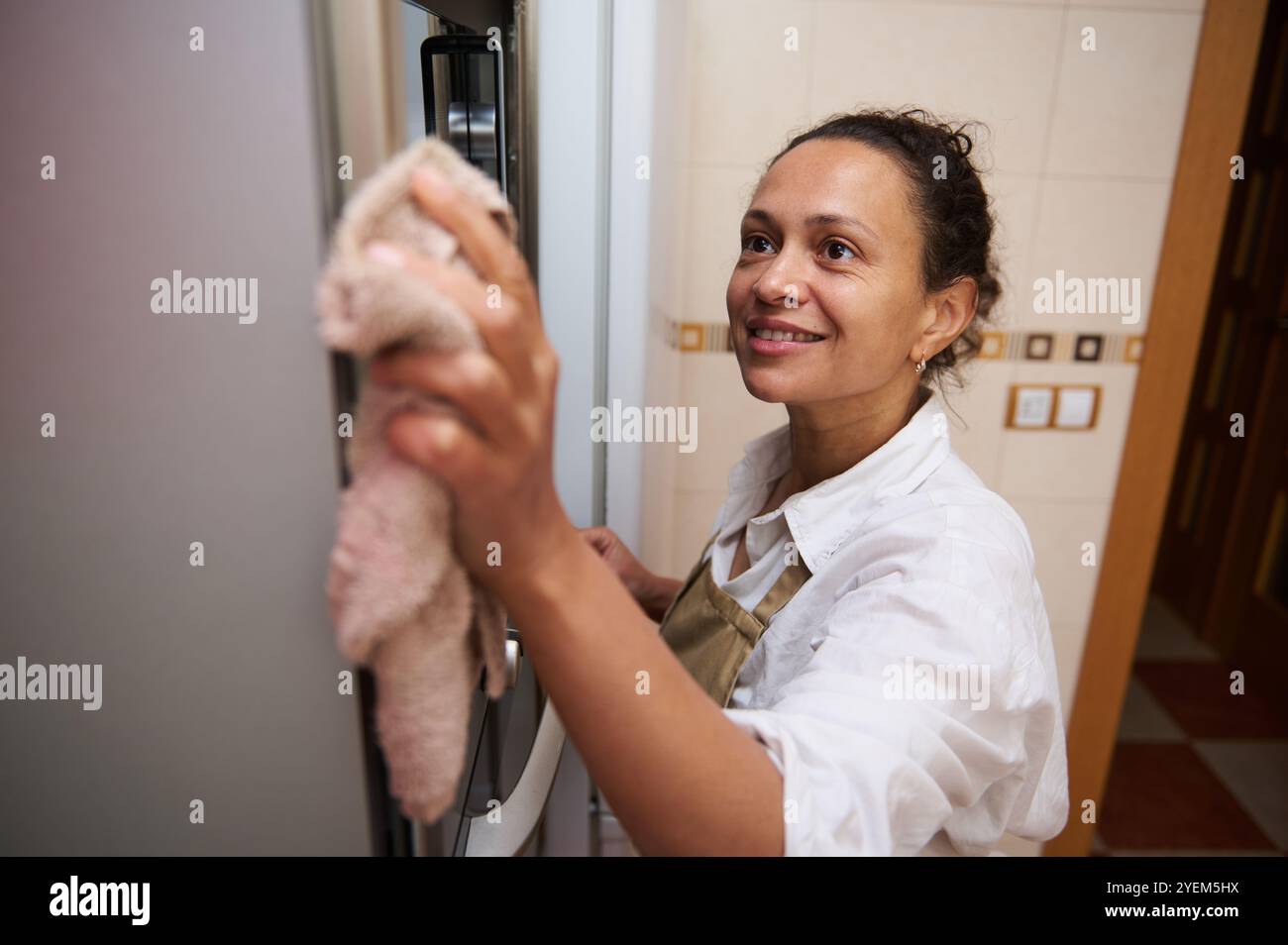 A cheerful woman wearing an apron cleans the kitchen, wiping down ...