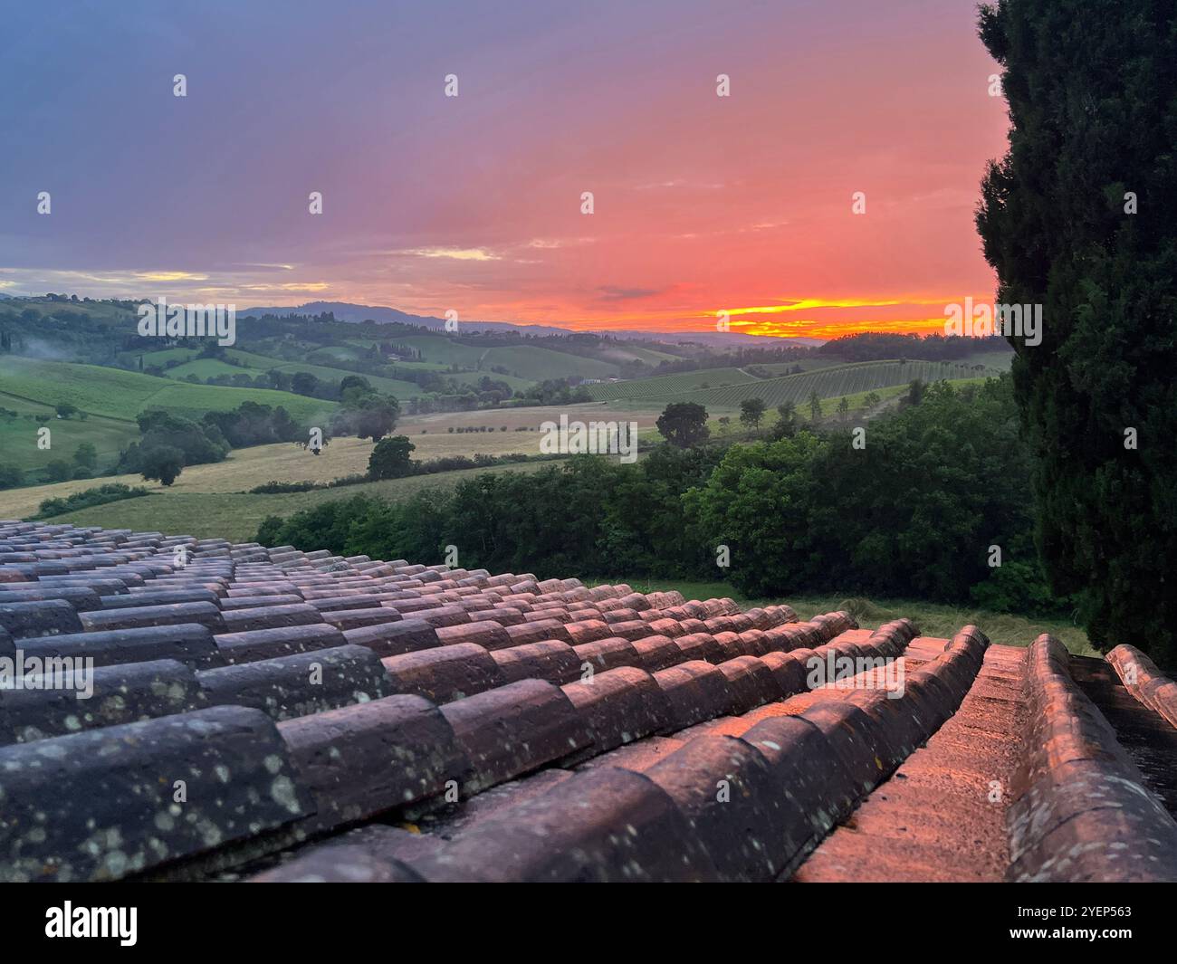 A beautiful sunset is shown over the rolling hills of Tuscany, Italy, with the orange sunlight reflecting on the tile roof in the foreground. - Smartphone Captured Stock Image
