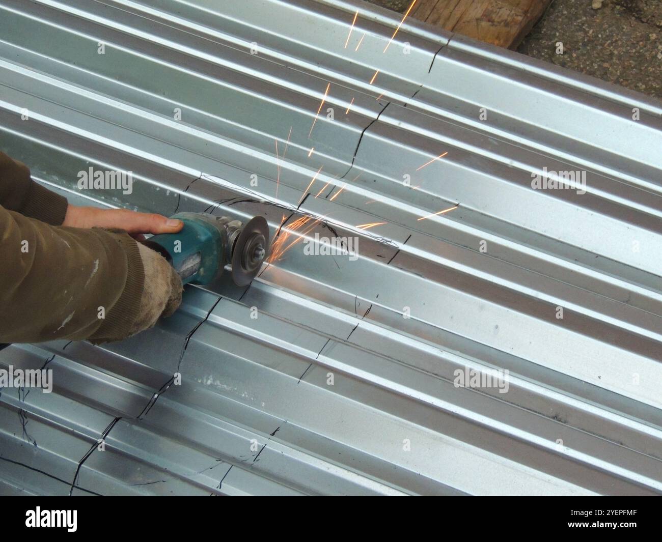 A man in dirty work clothes and gloves cuts a metal sheet with a grinder along the black markings on a corrugated surface, causing sparks to fly. Stock Photo