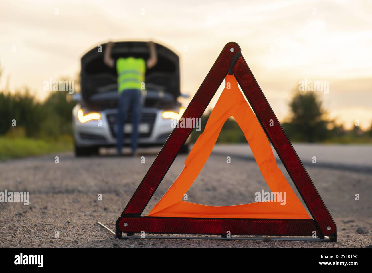 Unrecognizable sad driver in reflective vest. Male driver standing near broken car with open up hood. Red triangle to warn other road users of car bre Stock Photo