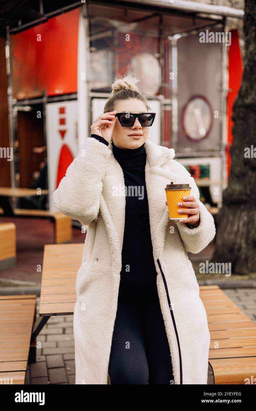 A young pretty woman in a white fur coat and sunglasses with coffee ...