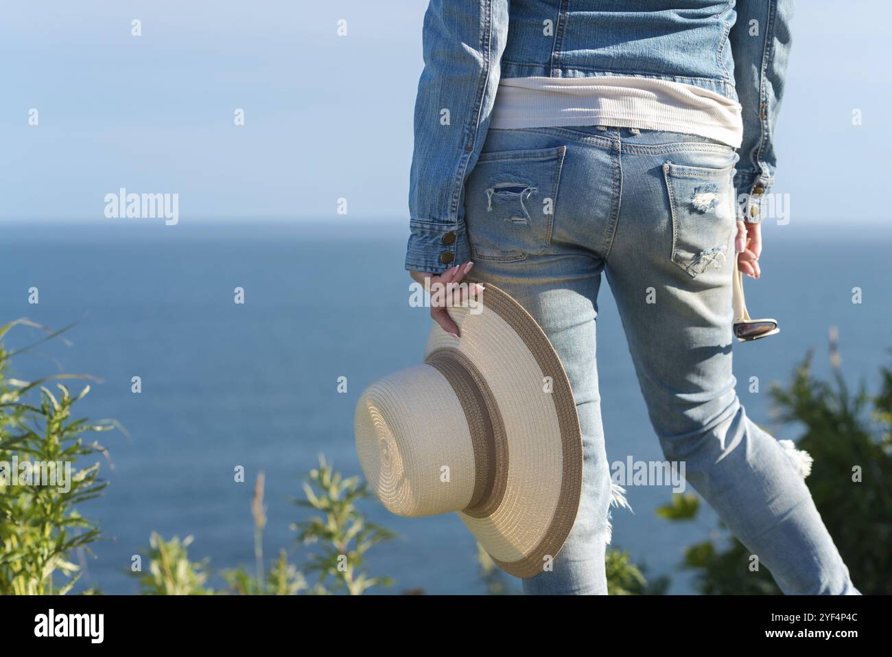 Cropped rear view below waist on legs of woman in blue jeans. Unrecognizable hipster female holding straw hat in one hand and sunglasses in other, sta Stock Photo