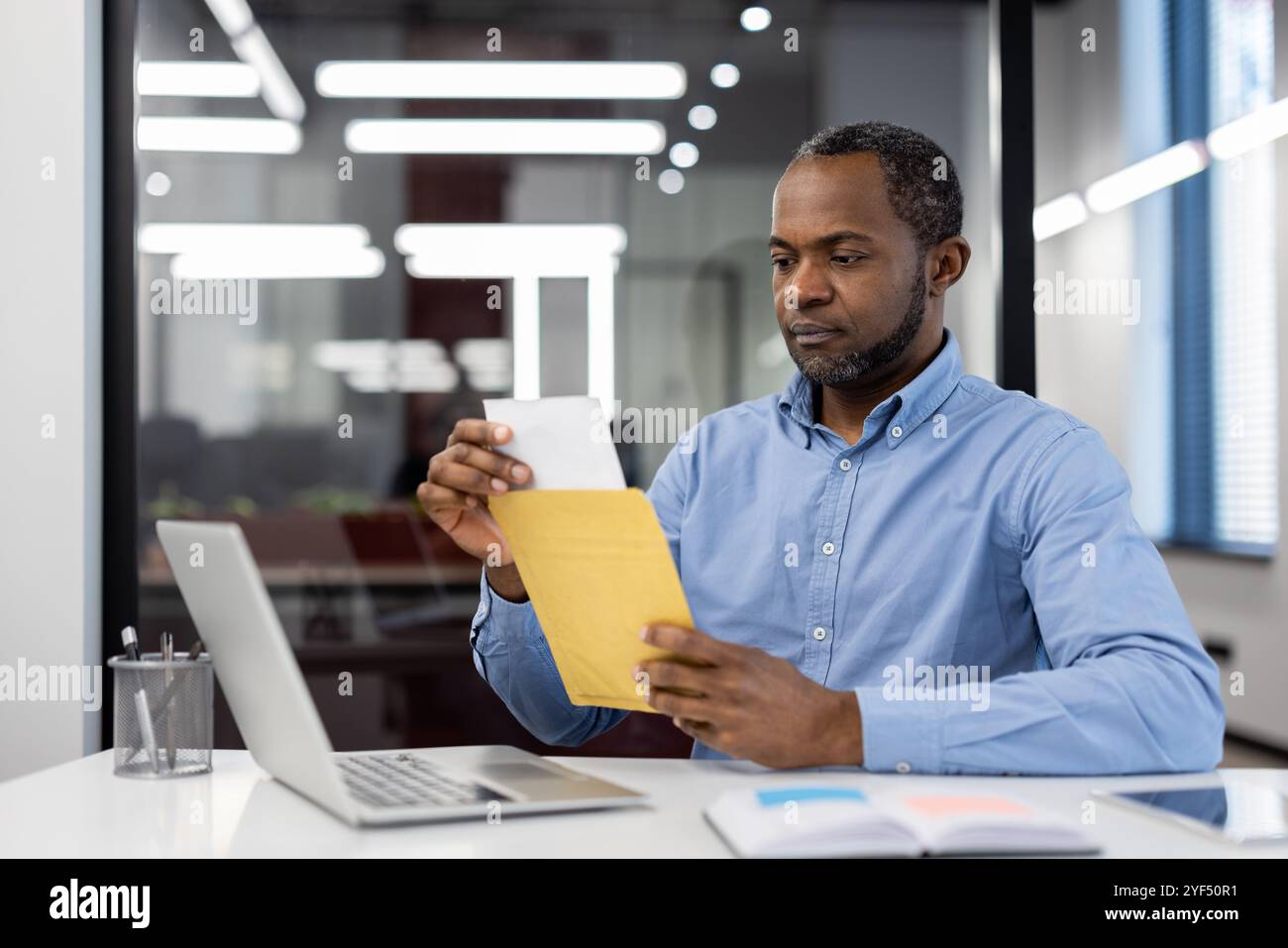 Mature African American businessman in office using laptop, opening envelope. Focused expression suggests concentration, professionalism. Bright lighting, modern setting denotes corporate environment Stock Photo