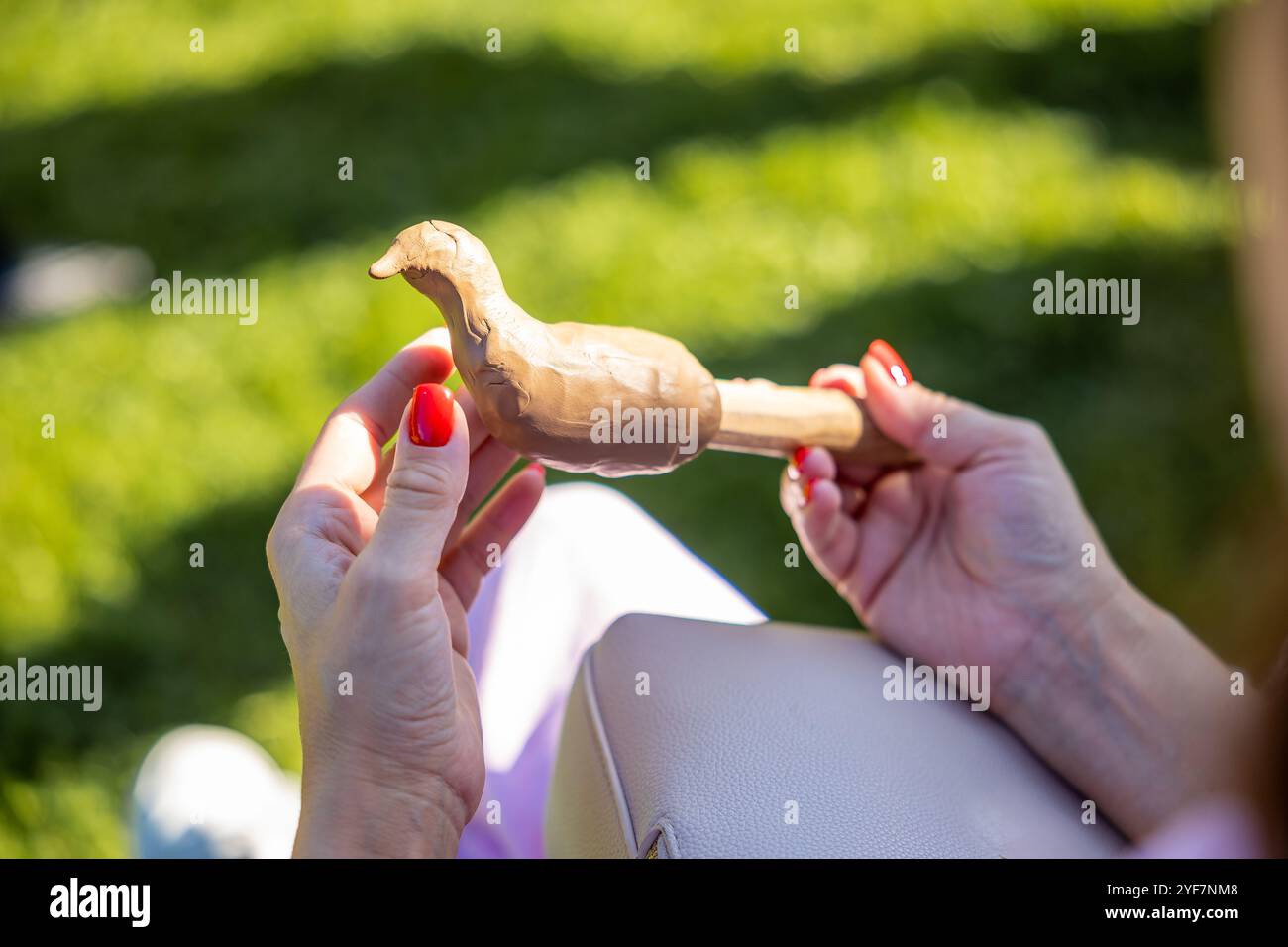 A person with red painted nails holds a wooden bird carving, possibly a duck, while sitting on grass. The green background contrasts with the vibrant Stock Photo