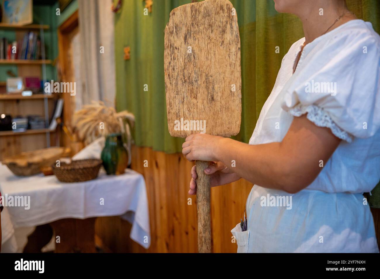 A person in a white garment holds a wooden paddle in a room with wooden paneling, a table with a basket, a green vase, and a bundle of wheat. Stock Photo