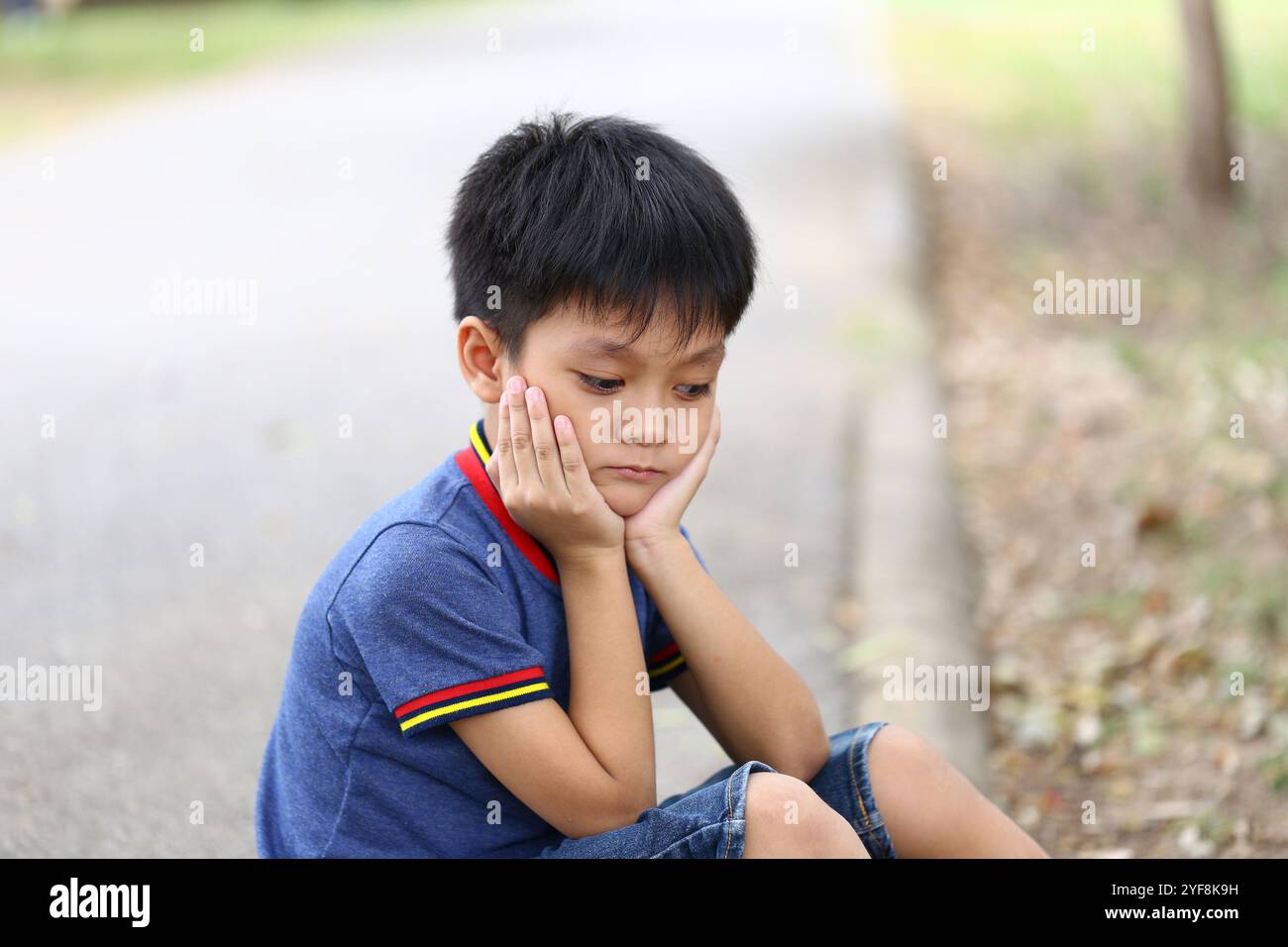 A young boy sits alone on a pathway, resting his chin on his hands ...
