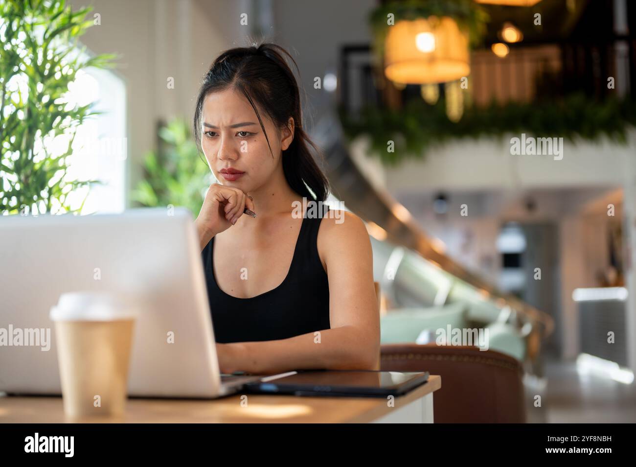 A stressed, thoughtful Asian woman working remotely from a coffee shop ...