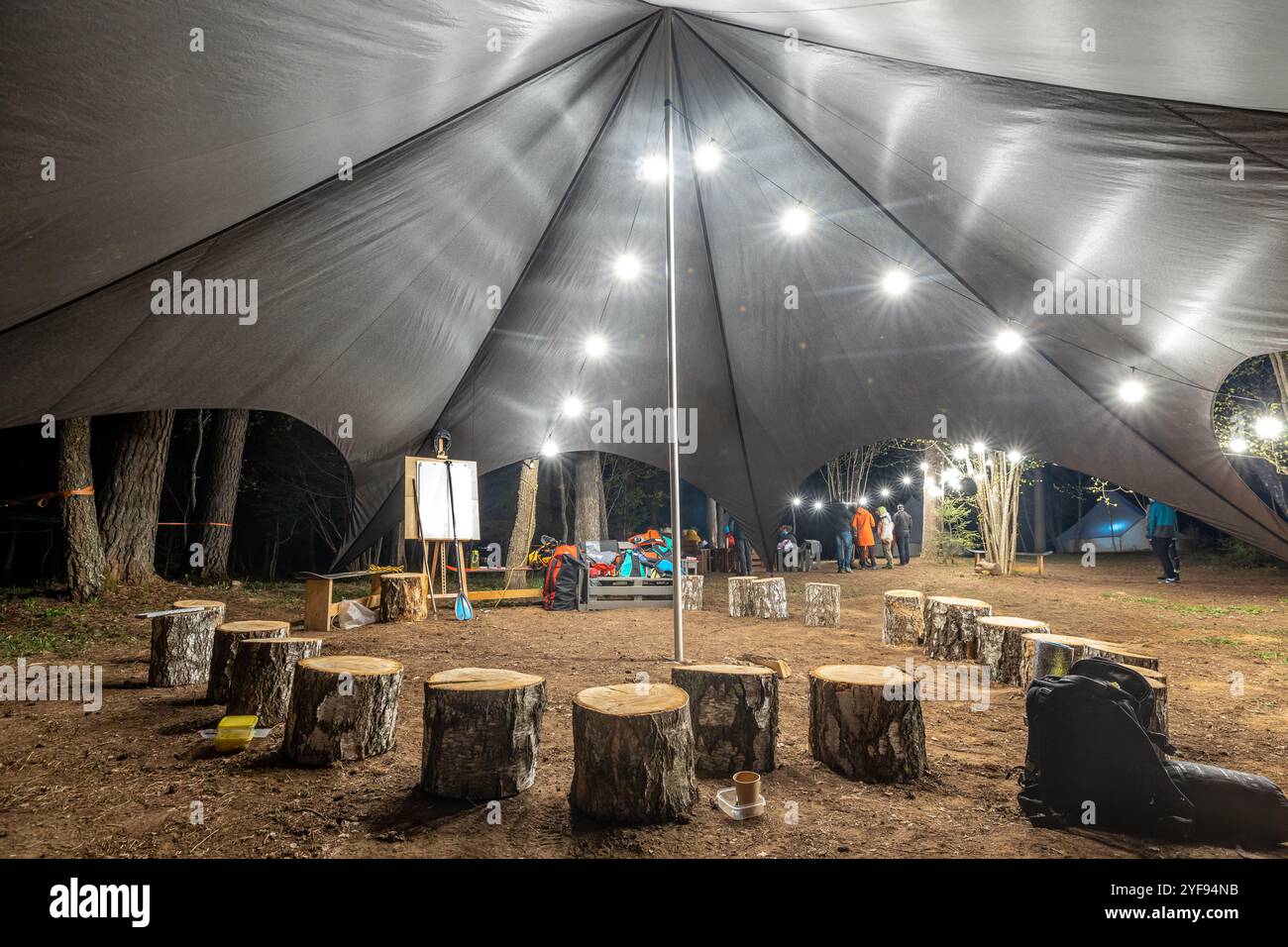 campers gathering under a spacious canopy tent at a forest campsite, illuminated by overhead lights for a cozy night-time retreat among the trees Stock Photo