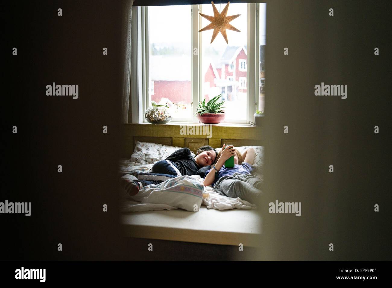 Smiling boy lying beside non-binary sibling using smart phone while lying on bed at home Stock Photo