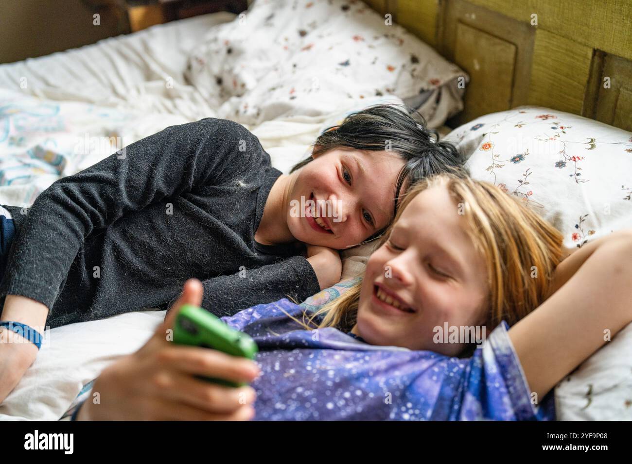 Excited boy lying beside non-binary sibling using smart phone while lying on bed at home Stock Photo