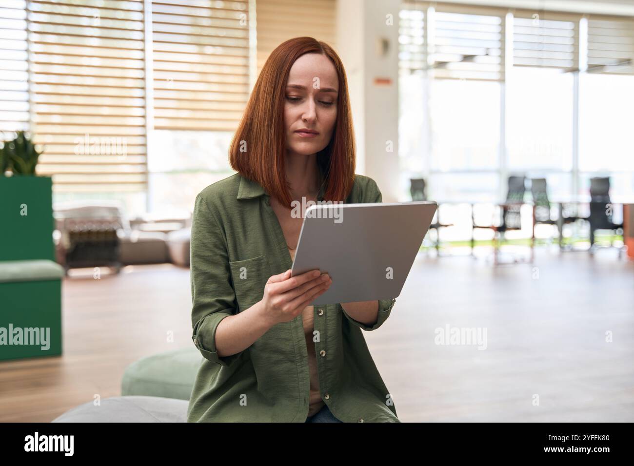 Young businesswoman is sitting in the lobby of her office, concentrating on reading something on her tablet Stock Photo