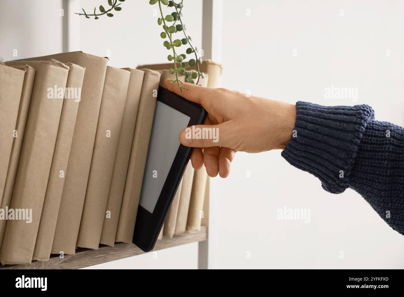 Young man taking e-reader from shelf with books at home, closeup Stock Photo