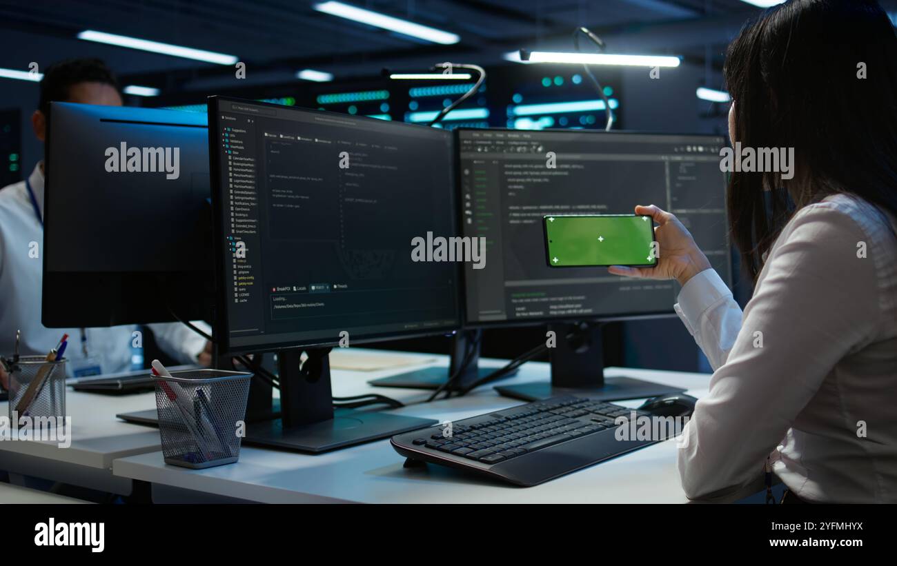 Zoom in on green screen mobile phone used by data center employee troubleshooting issues. Engineer uses mockup smartphone device in server hub, establishing performance benchmarks, camera B Stock Photo
