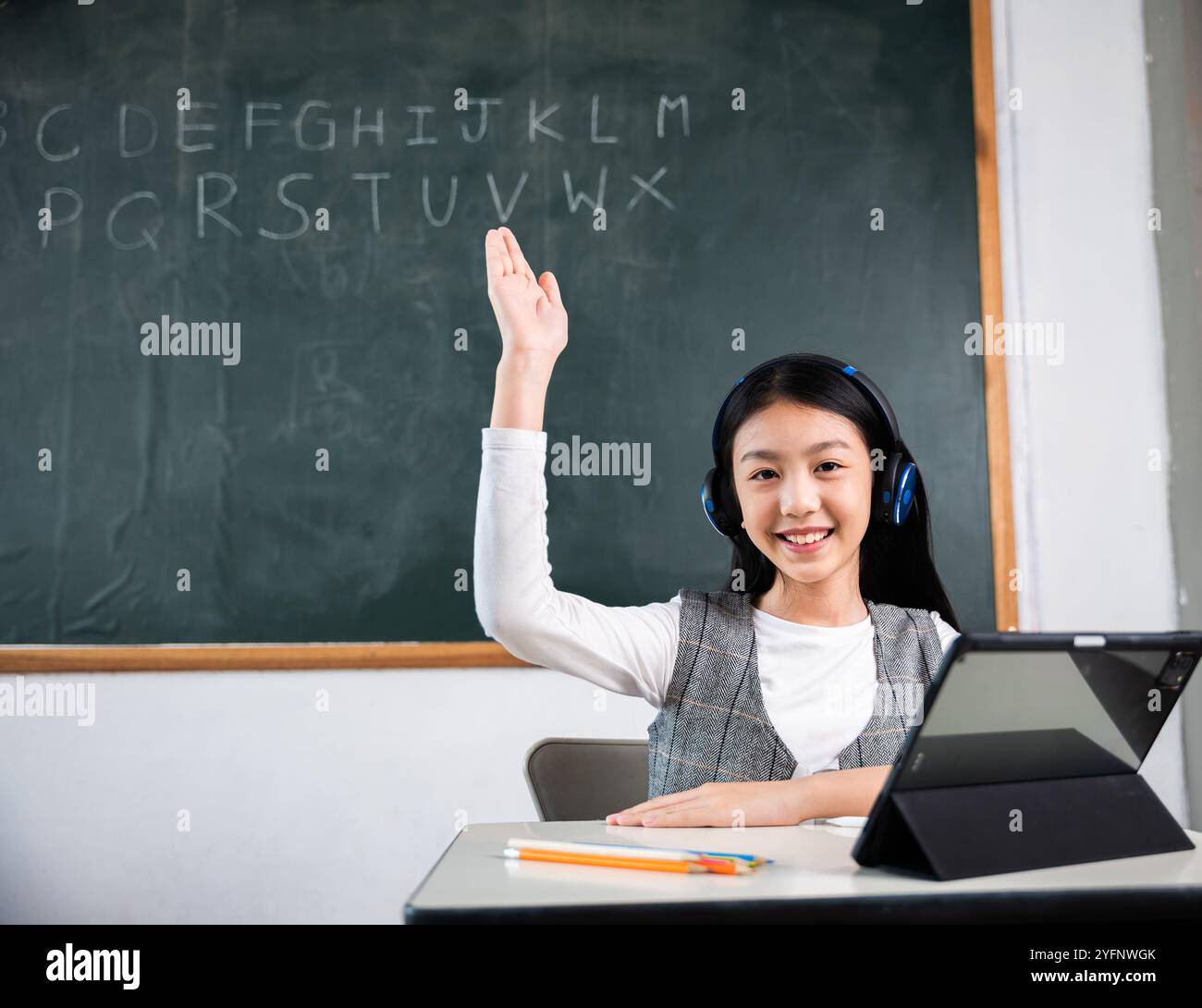 A young girl is sitting at a desk in a classroom, holding up her hand ...